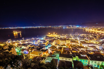Naklejka premium Illuminated old town of Nafplion in Greece with tiled roofs, small port, bourtzi castle, Palamidi fortress at night.