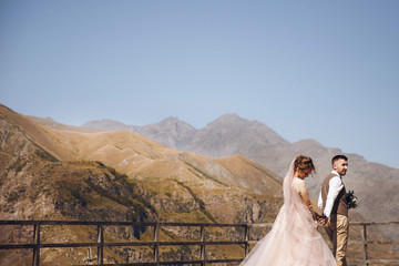 Groom in modern suit and bride in charming pink dress pose on the terasse with great mountain view...