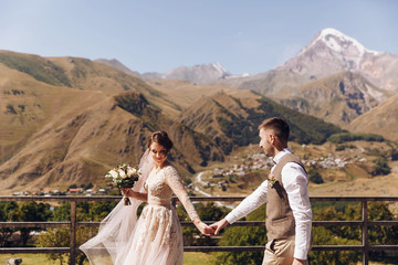 Groom in modern suit and bride in charming pink dress pose on the terasse with great mountain view...