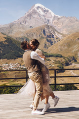 Groom in modern suit and bride in charming pink dress pose on the terasse with great mountain view...
