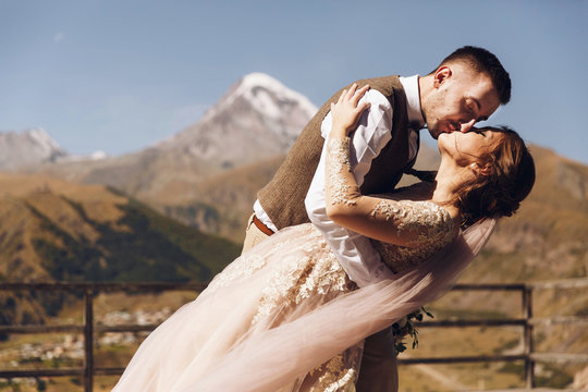 Groom in modern suit and bride in charming pink dress pose on the terasse with great mountain view in Georgia