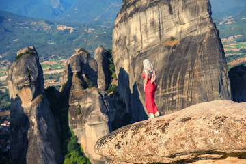 Alone girl in a red dress and scarf on the edge of the rock in Meteora. Creece in Thessaly. Pilgrimage in Greece