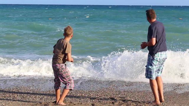 Father and son having fut at seaside during summer vacations. Adult man and young boy throwing stones into sea water cheerfully. Happy family concept.