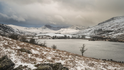 Beautiful Winter landscape image of Llynnau Mymbyr in Snowdonia National Park with snow capped mountains in background
