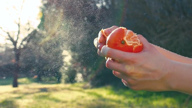 Hands of woman peel tangerine (mandarin) outdoor on background of spring meadow. Orange fruit splashes are fontaining to air. Slow motion, close-up.