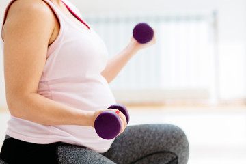 Fototapeta na wymiar Close up of pregnant woman sitting on fitness bal and working with dumbbells. Sport, maternity and liesure concept.