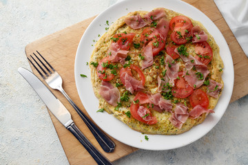 omelette with tomatoes, prosciutto ham and parsley garnish on a kitchen board, light gray background, high angle view from above, copy space