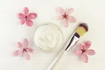 Obraz na płótnie Canvas Beauty skincare cream in cosmetic jar, delicate fresh pink spring blossom, application brush, top view white wooden table.