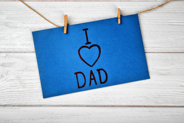 Card with phrase I LOVE DAD for Father's Day hanging on string against wooden background