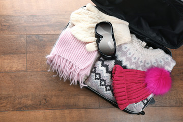 Open suitcase with warm clothes and ski goggles on wooden floor. Winter vacation concept