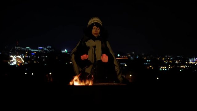 Hobo middle age woman warm at bonfire at cold windy night with far away city lights close-up