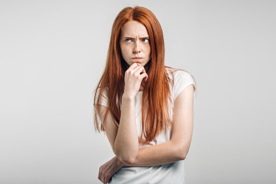 serious redhead young Caucasian woman holding arm on her chin and looking sideways with concentrated and focused expression