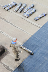 Worker installing roofing felt with heating and melting bitumen roll by torch on flame, high angle...