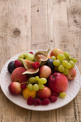 Top view on assortment of juicy fruits on white plate and wooden table background. Organic raspberries, apricots, apples, figs,plums, grapes - summer dessert or snack. healthy eating concept.