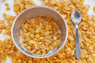 Cornflakes in a metal bowl with milk on a painted white wooden background. Cornflakes scattered on a wooden table with spoon.