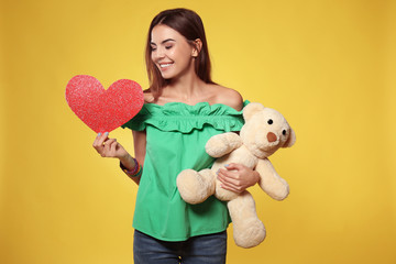 Fototapeta na wymiar Romantic young woman with paper heart and toy bear for Valentine's Day on color background