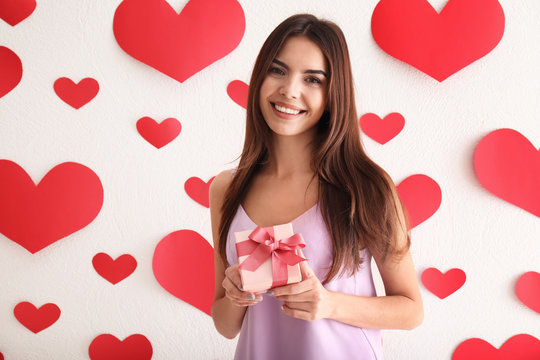 Beautiful young woman with gift for Valentine's Day near wall with paper hearts