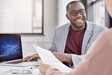Crew of financial mangers analyze financial report together, discuss solutions and share ideas. Glad smiling African American office worker shows results of his project, brainstroms with colleague