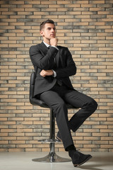 Handsome man in elegant suit sitting on chair near brick wall