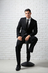 Handsome man in elegant suit sitting on chair near white brick wall