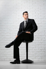 Handsome man in elegant suit sitting on chair near white brick wall
