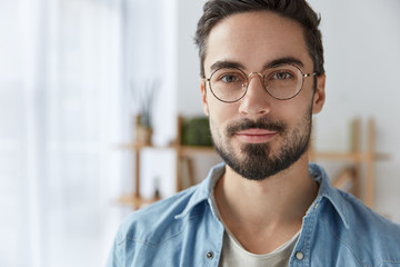 Close up portrait of handsome bearded man wears round spectacles, has appealing appearance with beard and mustache, dressed in fashionable clothes, stands against cozy interior. Student poses indoor