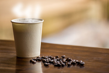 The paper cup of hot coffee with smoke and coffee beans on the wooden table in the restaurant with the feeling of retro oe vintage style.
