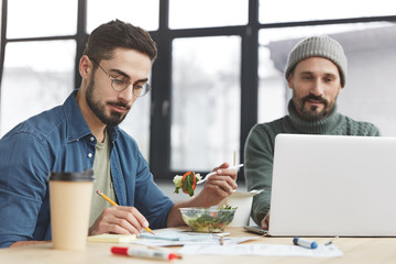 Concentrated handsome male managing expert writes down notes and eats fresh vegetable salad, fashionable bearded man works on laptop computer, collaboarte on common project. Dinner time at office