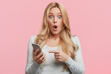Blonde young female with pleasant appearance looks with terrified expression in smart phone, reads...