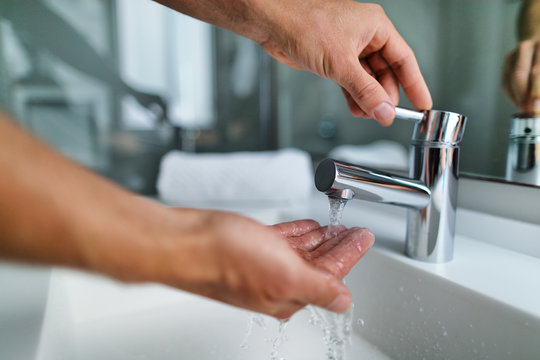 Man washing hands in bathroom sink at home checking temperature touching running water with hand. Closeup on fingers under hot water out of a faucet of a sink.