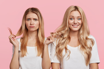 Envious young female compares her hair with friend, wants to have curls, discuss something, express different emotions, pose against pink background. Youth, friendship and relationship concept