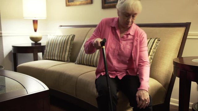 A Caucasian senior citizen woman with a cane enters the living room and sits on the edge of couch rubbing her arthritic knee which is causing pain.