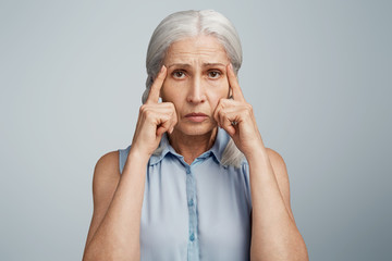 Displeased mature woman tries to concentrate on something, keeps fingers on temples, being dissatisfied and has headache after knitting, dressed in elegant blouse, isolated on blue background