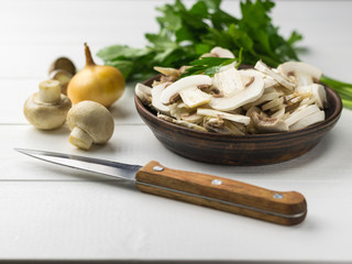 Fresh sliced mushrooms in a clay bowl and onion and herbs on a white wooden table.