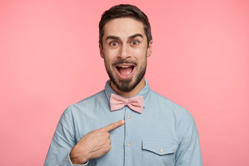 Puzzled young male model indicates at himself with indignant expression, demonstrates new bowtie, isolated over pink background. Handsome man looks in bewilderment at camera, gestures indoor