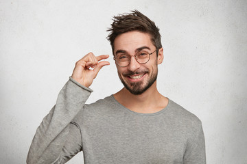 Positive handsome man with stylish hairdo, dressed in casual sweater, shows something very tiny or small, being in good mood, poses against white concrete background. Young fashionable male gestures - obrazy, fototapety, plakaty