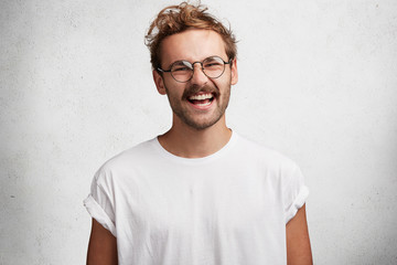 Positive pleased handsome stylih young man in round eyewear and casual t shirt, laughs at something...