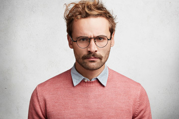 Fototapeta premium Horizontal shot of grumpy angry man frowns face in dissatisfaction, looks through spectacles, has gloomy expression, poses against white concrete background. Discontent displeased young human male