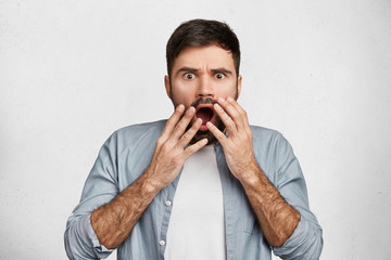 Frightened bearded male brunet covers widely opened mouth nervously, exclaims with anger, dressed...