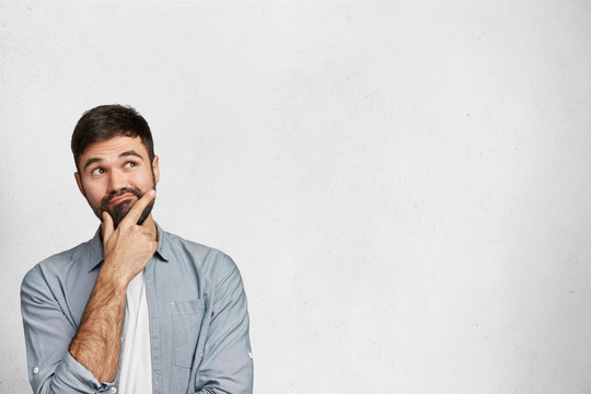 Indoor shot of handsome brunet male with pensive expression, dressed in fashionable shirt, tries to remember something, isolated over white background with copy space for your promotional text