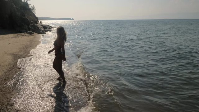 Girl runs and dances on the beach. She rushes into the frame, jumps, rejoices, enters the water, sits on the beach. The image is contrasting, almost silhouetted.