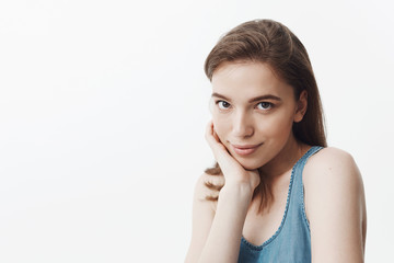 Fototapeta premium Close up isolated portrait of beautiful young caucasian female student with dark hair in blue shirt holding head with hand, looking in camera with gentle smile and relaxed face expression.