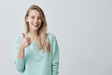 Happy young caucasian female wearing blue long sleeved shirt making thumb up sign and smiling...