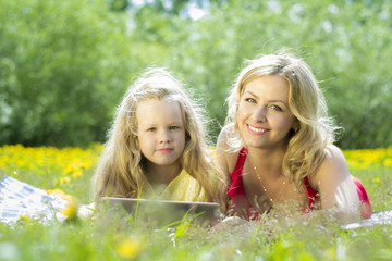 Beautiful woman and girl playing on a tablet, in the nature, lying on a blanket in the grass.
