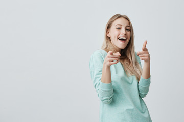 Happy positive joyful blonde woman with dyed hair smiling pleasantly pointing with index finger at you. Emotional smiling beautiful girl with wide opened mouth posing indoors. Advertising something.