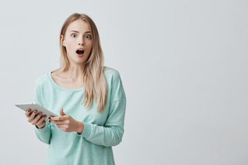Naklejka premium Isolated shot of shocked young blonde female wearing light blue sweater, looking and screaming with excitement and joy at the camera with tablet in hands. Modern technologies concept