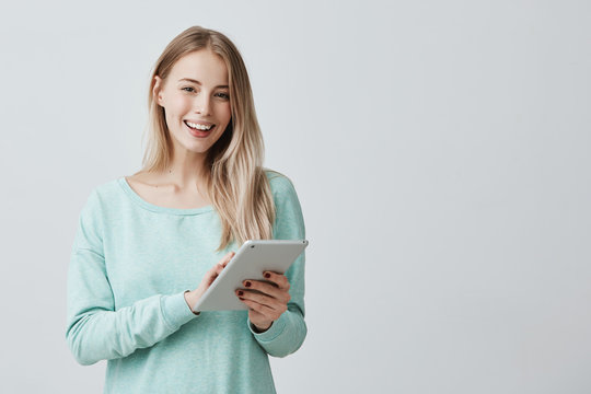 Beautiful female with long blonde hair using tablet for education or work at compilation of business charts. Smiling postive girl using modern technologies posing indoors.