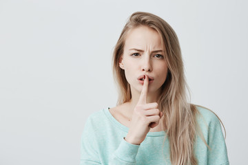 Naklejka premium Beautiful blonde female wears casual light blue long-sleeved shirt, shows hush sign, has serious expression, asks best friend not to tell secret anybody, hopes for sympathy and silence.