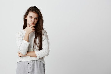 Beautiful girl with dark long hair, dark eyes, keeps hand on chin, looks pensively at camera, isolated against gray background. Confident thoughtful serious young female thinking about her next step