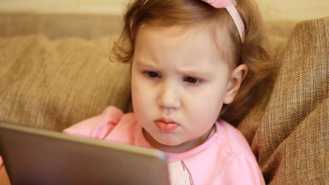 A little cute girl is sitting in sofa in a living room, looking cartoon and playing the game on a tablet.. Funny child looks on the computer screen and plays downloaded application. Close-up portrait